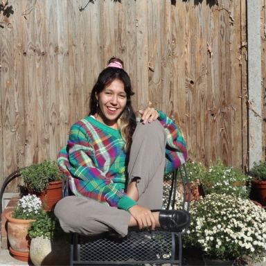 Photo of Alice Reid in her garden, sitting in a chair with her knee up, and smiling.