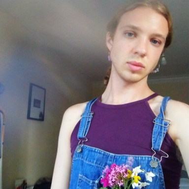 Selfie image of Harry Lindsey, wearing a purple top and denim dungarees with a small bouquet of flowers in the front pocket.