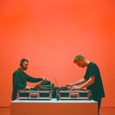 Bicep play electronic instruments facing each other in a red room