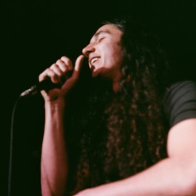 a man with long brown hair sings into a microphone and wears a black t shirt