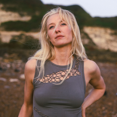 jessie has blonde hair and is posing on a beach with her hand on her hip. she is wearing a grey vest.