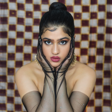 riya wears black sheer gloves and holds her face. the backdrop is chequered.