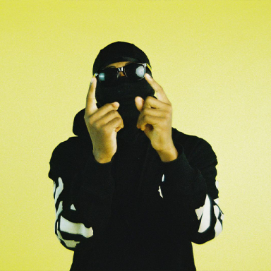 studiowyzz holds his hands up with index fingers up; he is wearing his signature balaclava with sunglasses and a black tracksuit