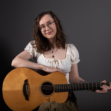 a woman with brown curly hair and glasses holds an acoustic guitar