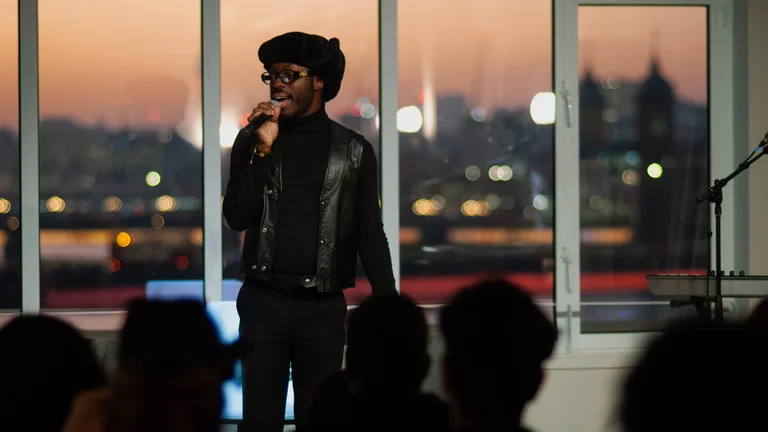 a person wearing glasses and an all-black outfit stands on stage, speaking into a microphone, with a sunset cityscape behind them