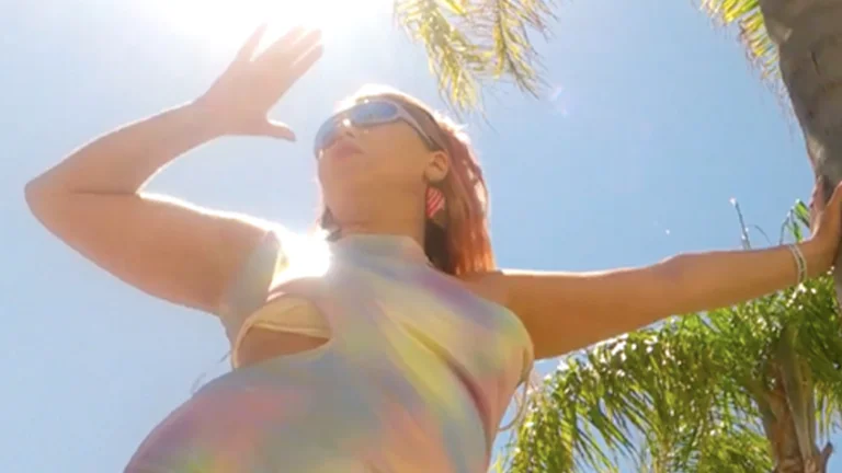 a woman is pictured from below, wearing a rainbow dress. she shields her face from the sun using her right hand