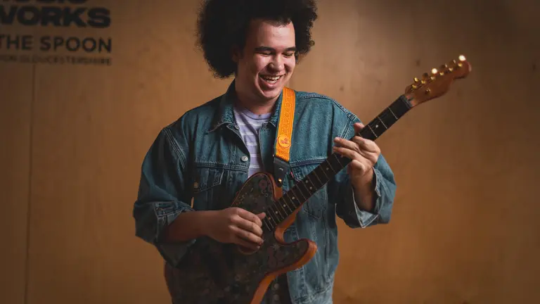 Image of Mackenzie playing the guitar standing up and smiling, wearing a denim jacket