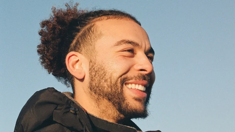 airz smiles, his hair in a bun. he wears a black jacket with a hood and is pictured against a blue sky