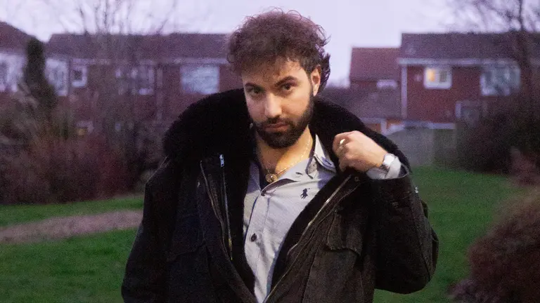 akam wears a blue shirt with a black hooded coat over the top. he is pictured in a park in front of some houses