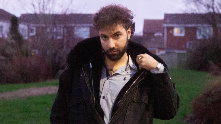 akam wears a blue shirt with a black hooded coat over the top. he is pictured in a park in front of some houses