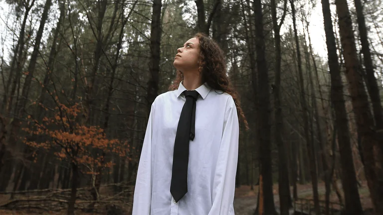 a woman with brown curly hair stands in a forest, wearing a white shirt and black tie