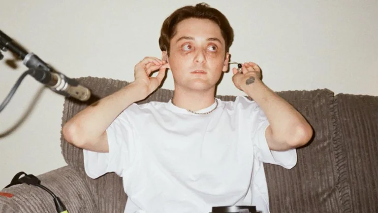 josh sits on a brown sofa and wears a white t-shirt. he checks his ear plugs