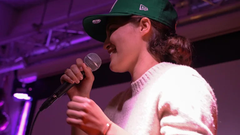 Iback wears a green cap and white jumper. she smiles and holds a mic