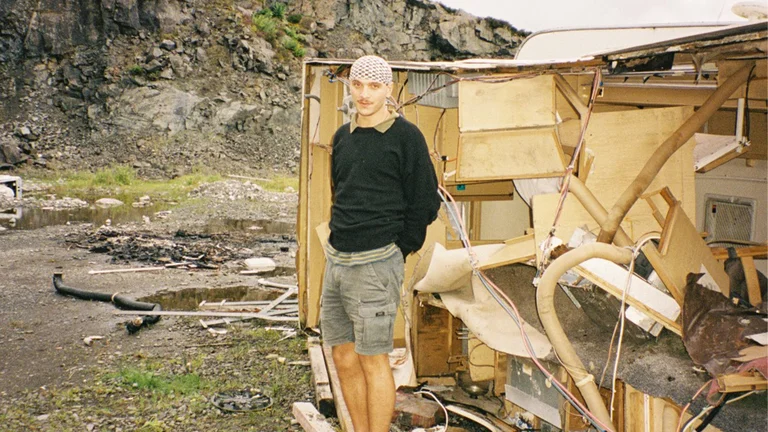 a person stands in a black jumper and grey shorts in front of a wooden wreckage