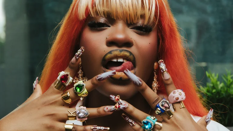 a woman with orange hair licks her lips and holds her hands up, laden with rings and long nails