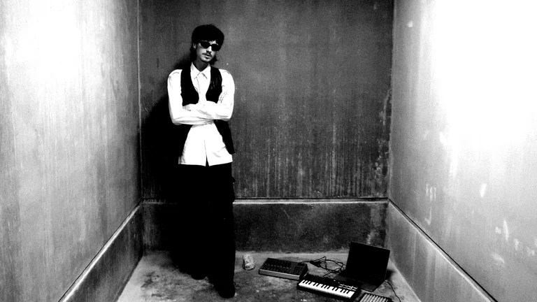 a man in a black waistcoat and trousers with sunglasses and a white shirt stands in a dingy room