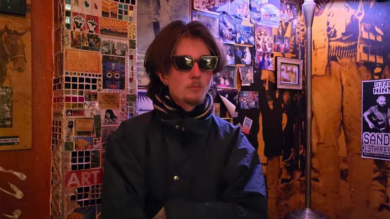 muckerhood poses in front of a wooden wall of photographs. his arms are folded and he wears a blue coat, sunglasses and striped scarf
