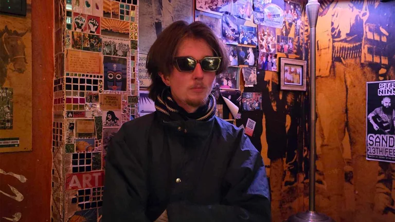 muckerhood poses in front of a wooden wall of photographs. his arms are folded and he wears a blue coat, sunglasses and striped scarf