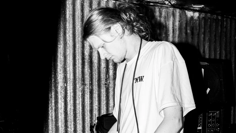 nailbreaker bows his head, his long hair tied up. he wears a white t shirt