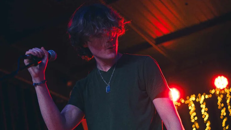 a man swishes his mop of fair hair on stage as he holds a standing mic