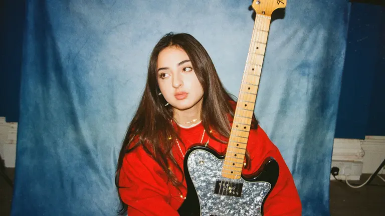 tala has long brown hair, a red jumper, and holds an electric guitar