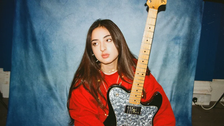 tala has long brown hair, a red jumper, and holds an electric guitar