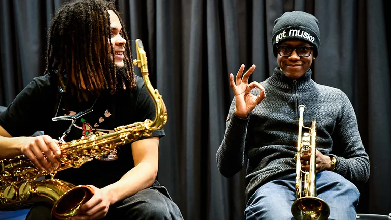 Two young people playing trumpet. One is making the OK hand gesture to the camera.