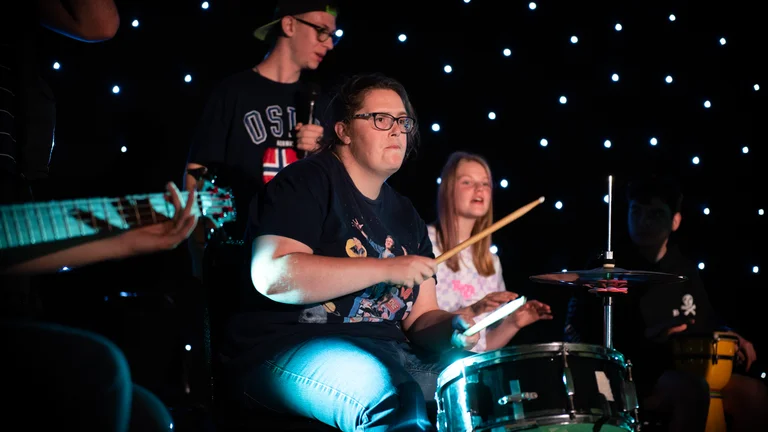 A young g person playing drums in a stage with two other people behind playing other instruments