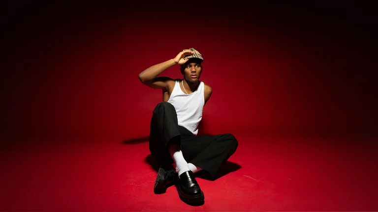exojay sits on the floor on a red backdrop with one hand on his cap