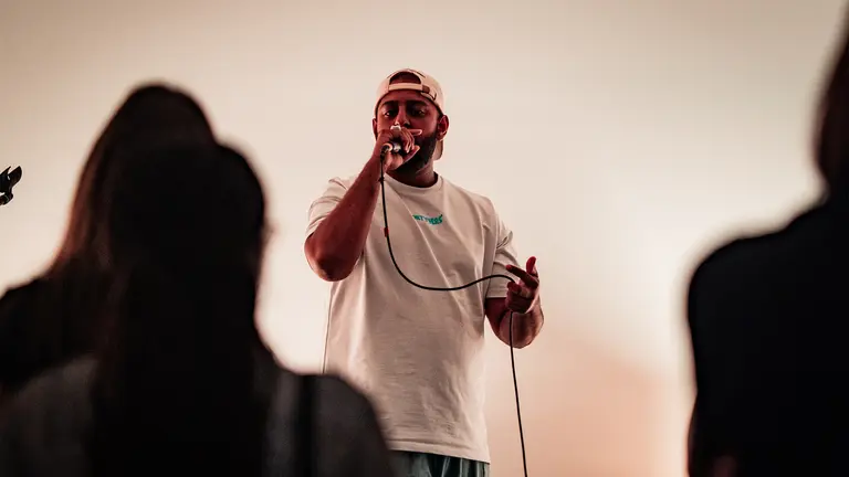 a man in a white t shirt wears a backwards cap and sings into a mic