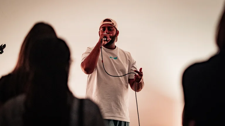 a man in a white t shirt wears a backwards cap and sings into a mic