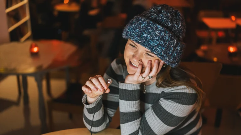 Young girl smiling in a stripey jumper and blue wooly hat. Resting her head on her hand at a table, being interviewed and holding a small mic
