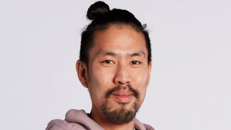 Dan Tsu headshot, white background, smiling, hair tied up in a bun, pink hoodie, arms folded. 