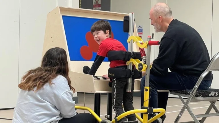 A young child in a red shirt uses a yellow mobility assistance device while being supported by two adults in what appears to be a therapy or rehabilitation setting. The child is smiling and appears engaged in the activity, with a blue backdrop visible behind them.