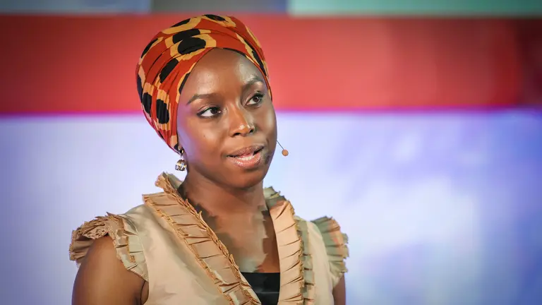 Author Chimamanda Adichie with a headworn microphone and a red and white background