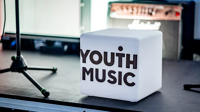 image of Youth Music branded light box. The light box is white with 'Youth Music' in black text. It is on a stage next to the feet of a microphone and in front of an amp