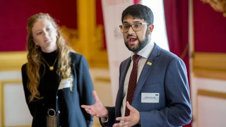 two young people standing young people. One is talking to an unseen crowd and the second person is looking at them. 