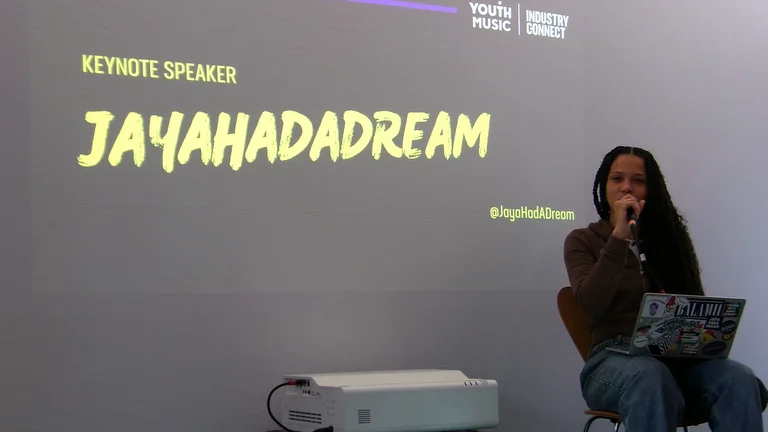 a woman with long braids sits on a chair on stage, holding a laptop and speaking into a microphone. the words 'keynote speaker: jayahadadream' is on the screen behind her.