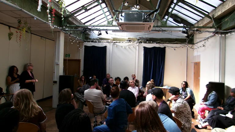 A diverse group of people seated in a circle engages in discussion in a bright room with high ceilings and large windows. The atmosphere is relaxed and focused.