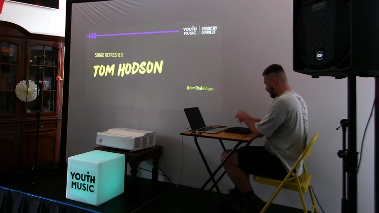 Man sits on a yellow chair playing music on a laptop on a small stage. A screen displays "Tom Hodson, Sonic Refresher," with Youth Music branding.
