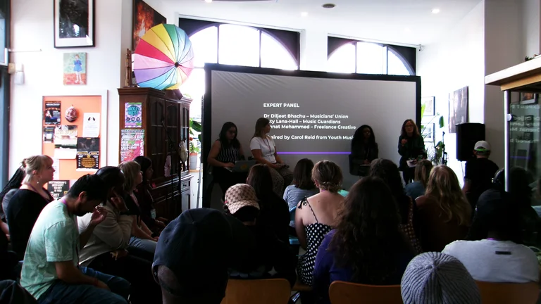 A diverse audience is seated in a cozy room, attentively watching an expert panel discussion projected onto a screen, with colourful decor around.