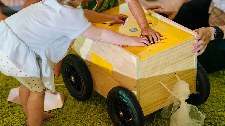 A child playing with a yellow toy. They and other children are touching the surface and making different sounds. 
