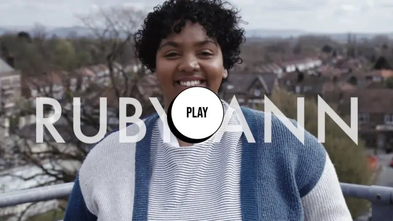 A person with a blue and white jumper over a city background. Their name Rubby-Ann appears and screen, together with a play button.