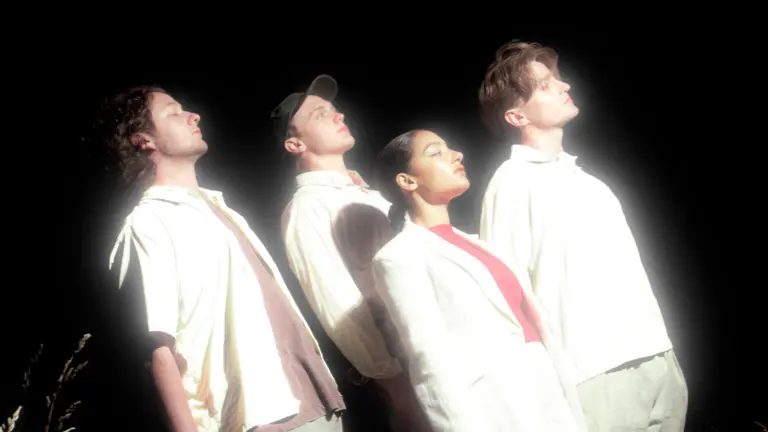Four members of English Teacher stand in grass at night, all looking upward with dramatic white lighting illuminating their faces against a black background. They wear white and cream clothing, with the woman in center wearing red boots.