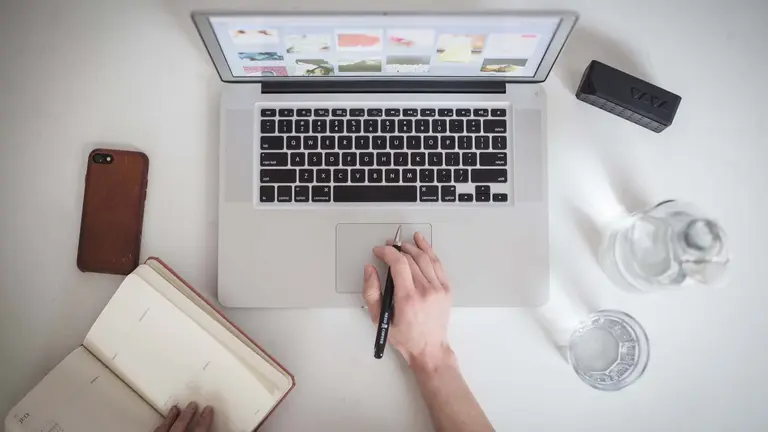 A person with a computer, only the hands are seen. One hand is on the touchpad and the other one is on a notebook