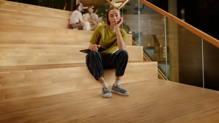 Person sitting down in wooden stairs. They're holding their chin with hand and looking at the camera. 