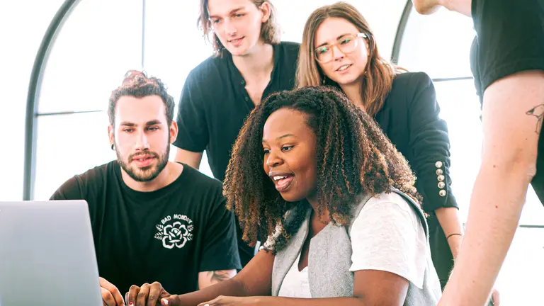 A Black woman in a computer with four colleagues behind, three standing up and another one sitting at her right. 