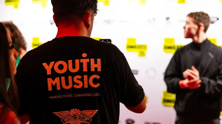 a youth music nextgen who is helping with the youth music awards is taking a picture of someone on the red carpet, we see his 'youth music' t-shirt in the foreground of the image