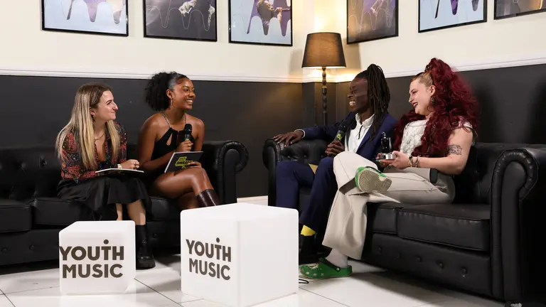 The youth music awards backstage presenters conducting an interview on black sofas with YMAs winners, in front of two lightboxes saying 'youth music'