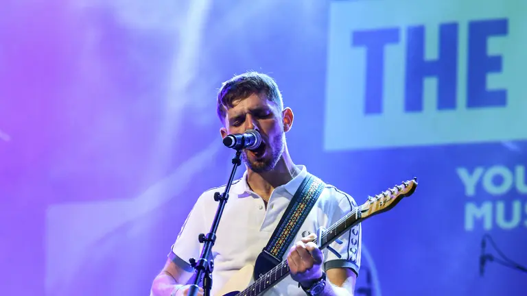 member of The 113 performing at the YMAs2025, singing into the mic and playing guitar
